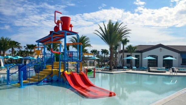 Un parque acuático con un tobogán rojo, estructuras de juego azules y amarillas, y una zona de piscina con sillones y sombrillas.
