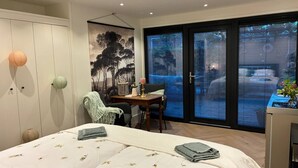 A bedroom with a bed, a desk, and a chair. There is a window with blinds, a door, and a wall with a painting.