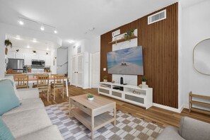 A modern living room with a TV, wooden panel wall, and a dining area with a kitchen in the background. - Family Comfort House / Sleeps 6  - 31-106 (Kissimmee)