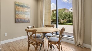 Uma sala de jantar com uma mesa de madeira redonda e seis cadeiras, um grande quadro abstrato na parede, e vista para um edifício e árvores através de grandes janelas.