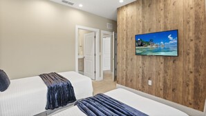 Quarto de hotel com duas camas, parede de destaque em madeira e uma televisão exibindo uma praia tropical.
