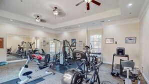 Ein Fitnessraum mit verschiedenen Trainingsgeräten, einem Deckenventilator und großen Fenstern.
