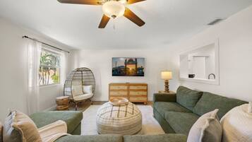 Un salon avec un ventilateur de plafond, un canapé vert, un pouf rond, une commode en bois et un tableau accroché au mur.