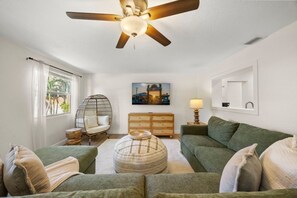 A living room with a ceiling fan, a green sofa, a round ottoman, a wooden dresser, and a wall-mounted picture. - Cocoa Beach Oasis: Boho Retreat w/ Huge Yard & Hammock - Sleeps 6 - 125 WG (Cocoa Beach)