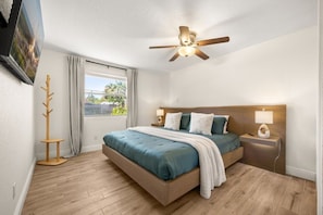 Un dormitorio con cama, ventilador de techo, mesita de noche, lámpara, perchero y ventana con cortinas.