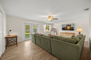 A living room with a sectional sofa, a ceiling fan, a lamp, and a painting on the wall. - Cocoa Beach Oasis: Boho Retreat w/ Huge Yard & Hammock - Sleeps 6 - 125 WG (Cocoa Beach)