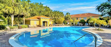 Una piscina con borde de baldosas, rodeada de palmeras y un edificio con techo de tejas rojas.