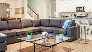 Un salon moderne avec un canapé d’angle, une table basse en verre et une cuisine en arrière-plan.