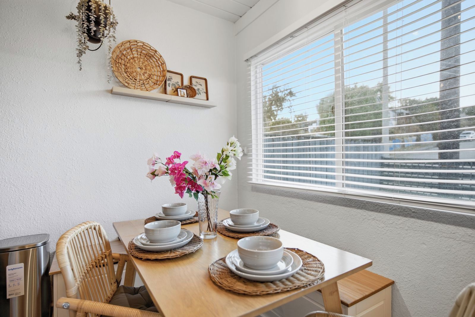 Un coin repas avec une table en bois dressée pour quatre personnes, des assiettes blanches et un vase rempli de fleurs roses.