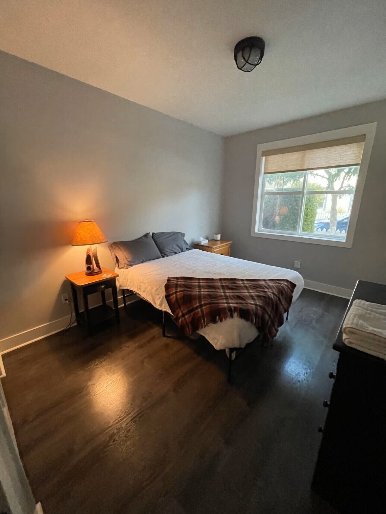 A bedroom with a bed, a nightstand, a lamp, a window, and a ceiling light.