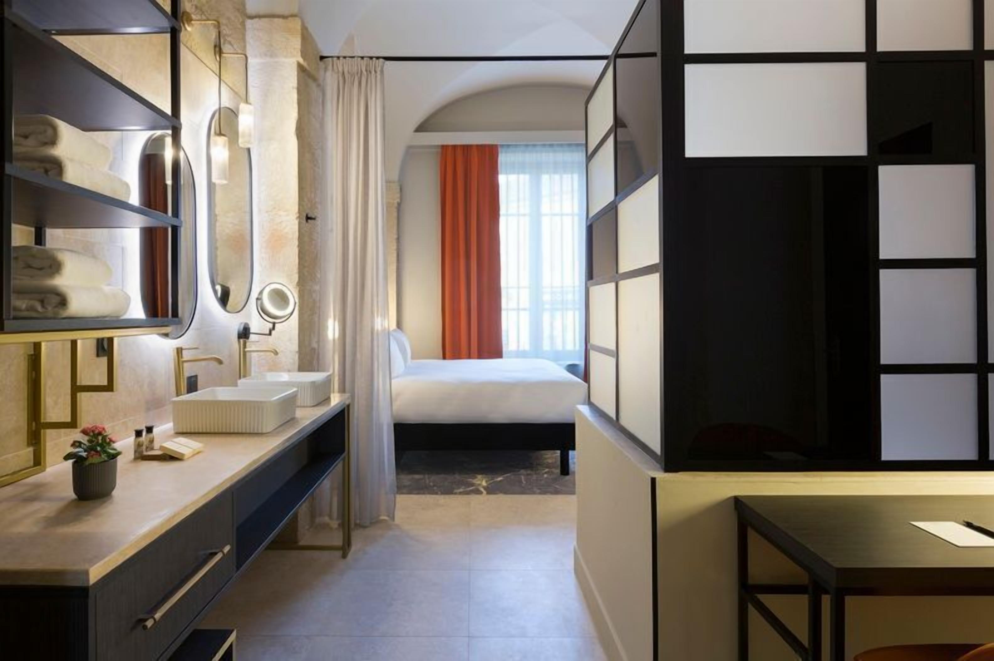 A modern hotel room with a large bed, a vanity with a mirror, a bedside table, and a view of the city.
