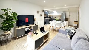 Una sala de estar moderna con un televisor de pantalla plana, un sofá gris, una zona de comedor con una mesa y sillas blancas, y una cocina al fondo.