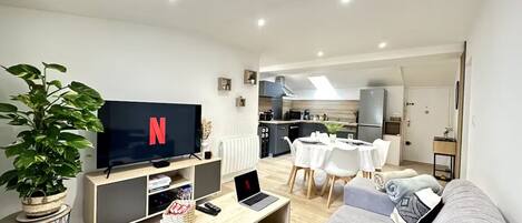 Ett modernt vardagsrum med en platt-TV, en grå soffa, en matplats med ett vitt bord och stolar, och ett kök i bakgrunden.