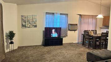 A living room with a television, a dining table, chairs, a plant, and artwork on the walls.