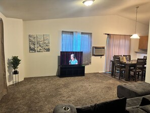 Una sala de estar con televisor, una mesa de comedor, sillas, una planta y obras de arte en las paredes.