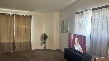 A living room with a large flat-screen TV, a black entertainment unit, a potted plant, and a window with sheer curtains.