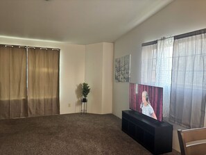 Un salón con un televisor de pantalla plana grande, un mueble de entretenimiento negro, una planta en maceta y una ventana con cortinas translúcidas.