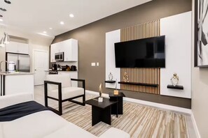 Ein modernes Wohnzimmer mit einer Couch, einem Couchtisch, einem an der Wand montierten Fernseher und einer Küchenzeile im Hintergrund.