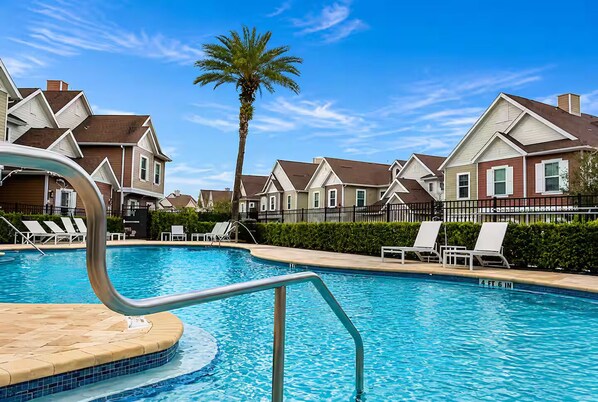 Una piscina con sillas de descanso, una palmera y casas adosadas al fondo.