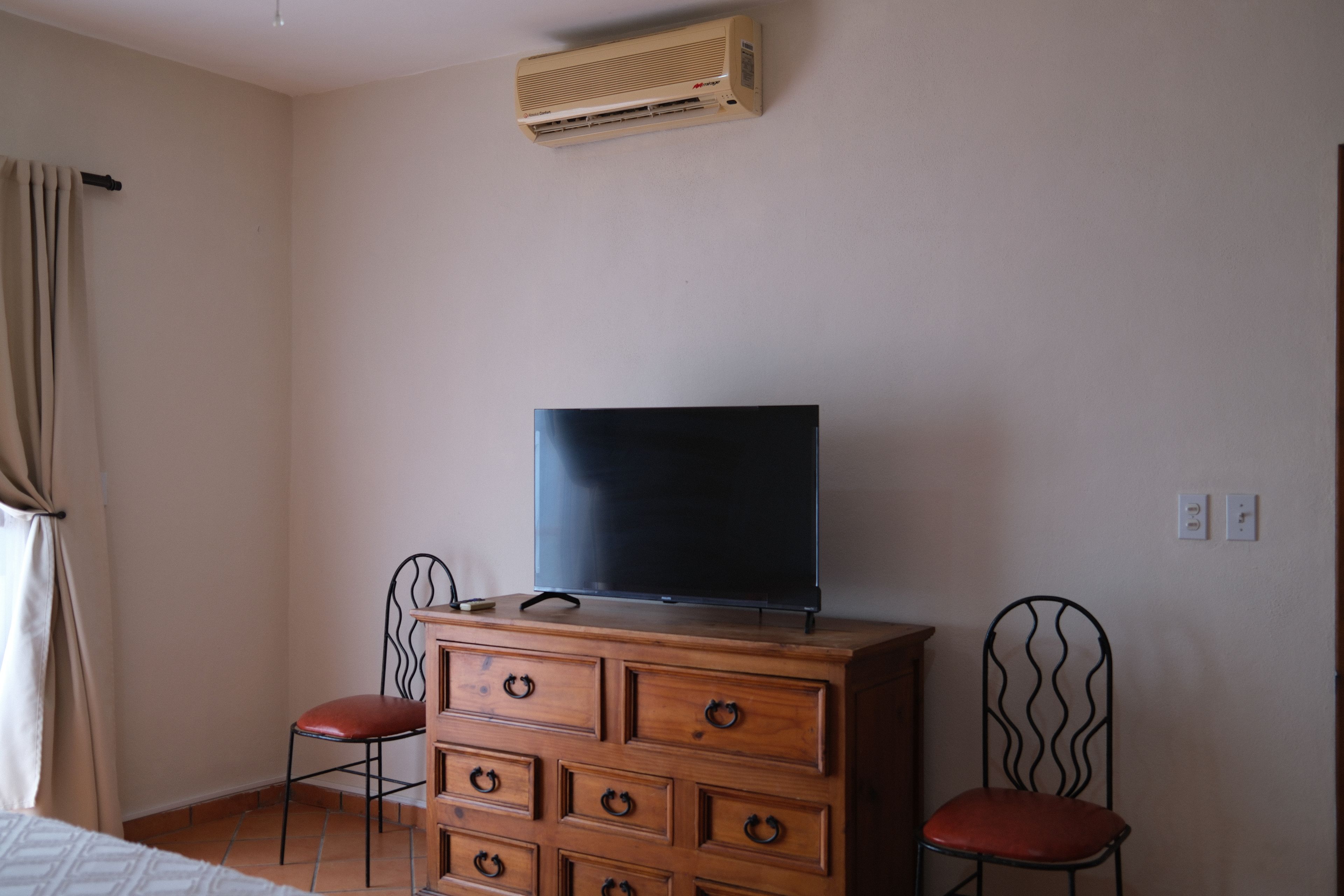 Una habitación con un aparador de madera, un televisor encima, dos sillas, una ventana con cortinas y un equipo de aire acondicionado en la pared.