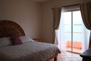 Une chambre avec un grand lit, une tête de lit en bois, une petite table de chevet et une vue sur la mer, visible à travers une porte-fenêtre coulissante.