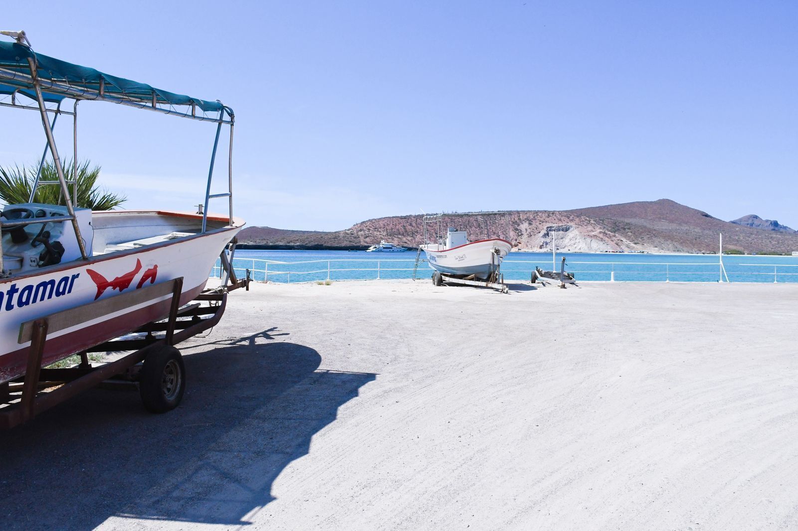 Un bateau nommé « Tamar » est posé sur une remorque, un deuxième bateau se trouve sur la plage, et un ciel bleu dégagé surplombe un paysage vallonné.