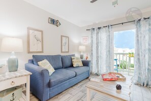 Ein Zimmer mit einem blauen Sofa, zwei Kissen, einem Tisch mit einem Buch und einer Fernbedienung, einer Lampe und einem Fenster mit Vorhängen.