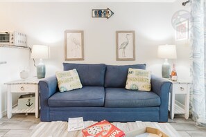 Ein Wohnzimmer mit einem blauen Sofa, zwei gerahmten Wandbildern, einem Beistelltisch mit Lampe und einem kleinen Tisch mit einem Buch und einer Flasche.