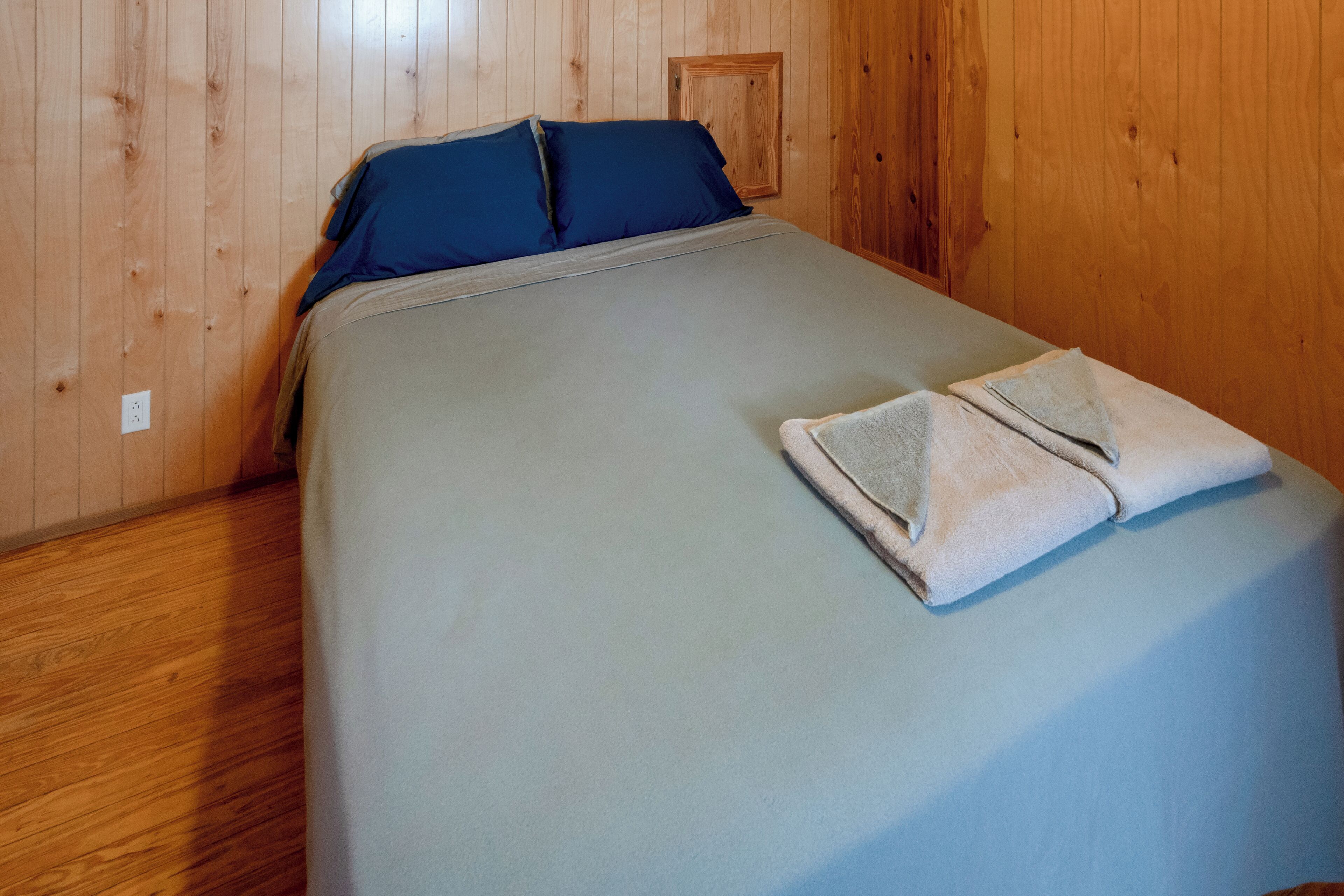 Una cama individual con sábana gris y dos almohadas, una toalla doblada y paredes de paneles de madera.