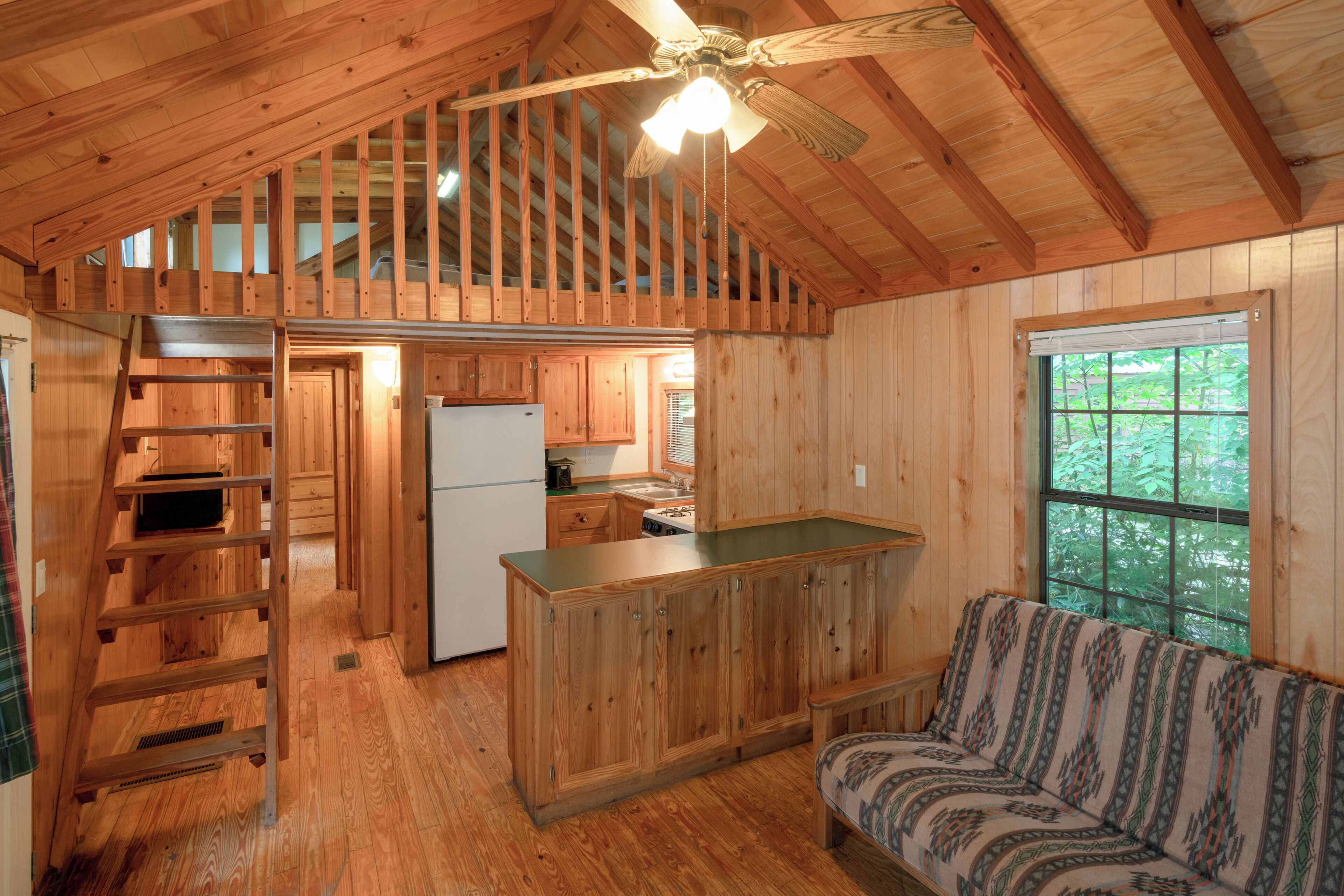 A cabin interior with a loft bedroom, a kitchenette, a sofa, and a large window.