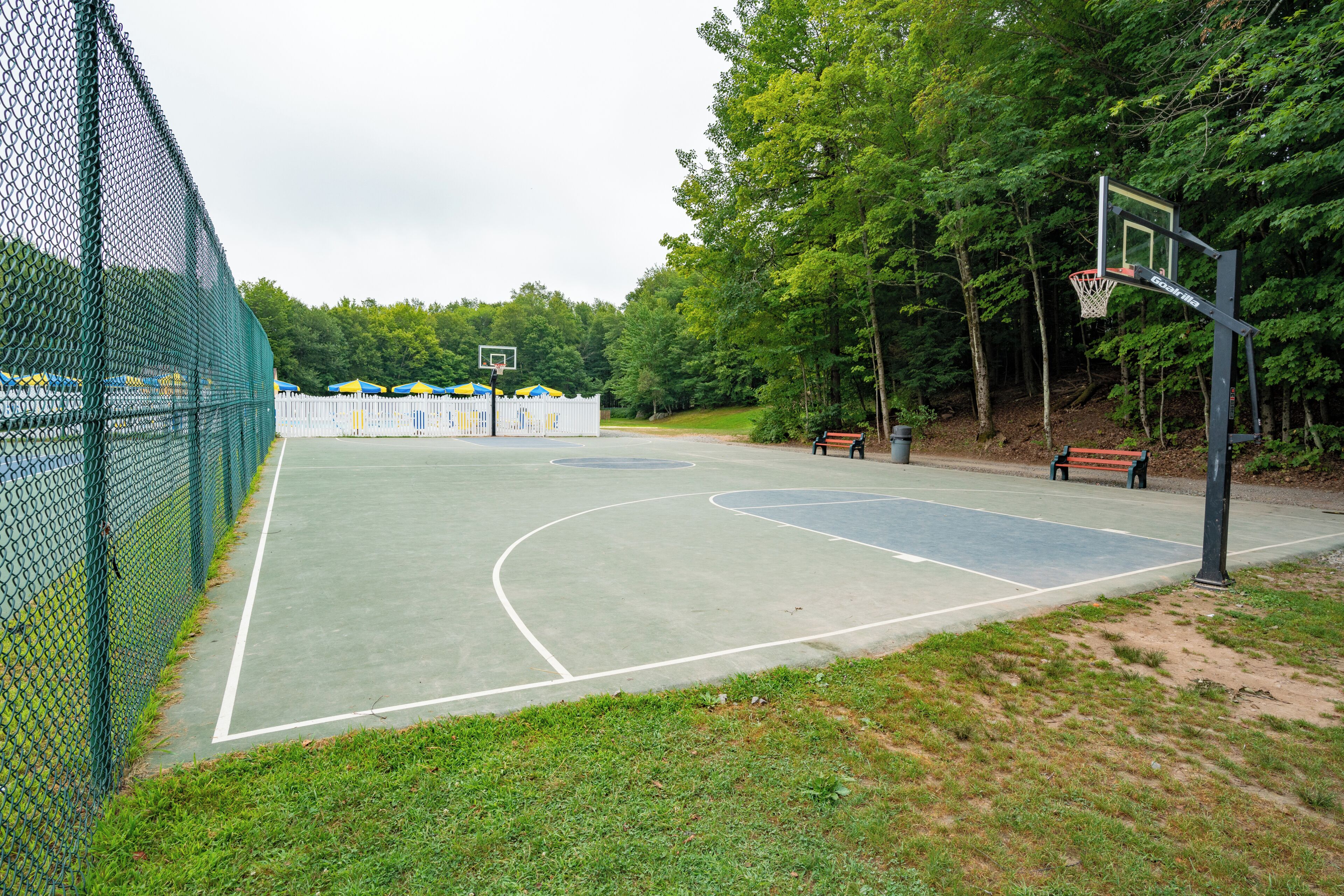 An outdoor basketball court with a chain-link fence, a basketball hoop, and a white picket fence in the background.