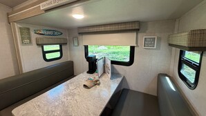 Interni di un camper compatto con zona pranzo, macchina del caffè e finestra vista su del verde.