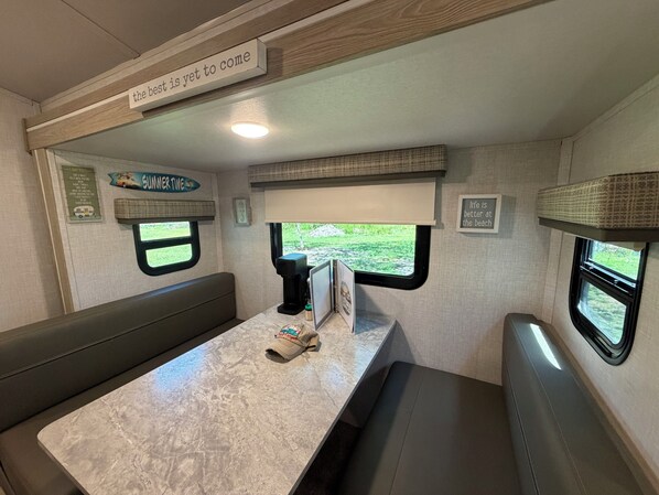 Een compact interieur van een camper, met een eethoek, een koffiezetapparaat en een raam met uitzicht op groen.