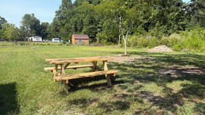 Un tavolo da picnic in legno in un prato erboso, sullo sfondo alberi e un piccolo edificio.