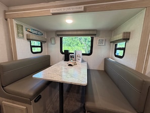 Ein gemütliches Wohnmobil-Interieur mit Tisch und zwei Sitzgruppen, einem Fenster mit Blick auf Grünflächen sowie motivierenden Wandbildern.