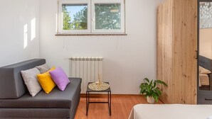 Een kamer met een grijze bank, een tafeltje met een decoratief object, een radiator, een houten garderobe en een raam met uitzicht op bomen.