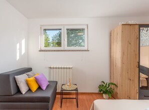 Ein Zimmer mit grauem Sofa, einem kleinen Tisch mit einem dekorativen Gegenstand, einer Heizung, einem Holzschrank und einem Fenster mit Blick auf Bäume.