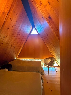 An attic bedroom with wooden walls, a triangular skylight, two beds, and a wicker chair. - Ger's Chalet (Devesa de Saga)