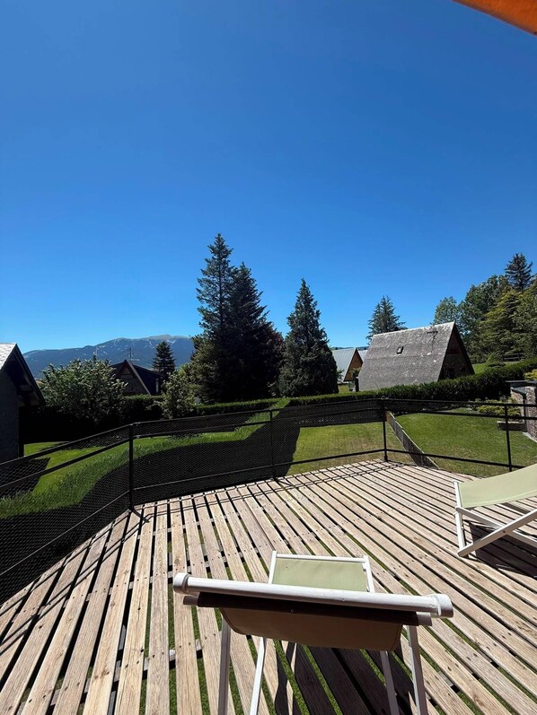 A wooden deck with a chair, a netted fence, and houses in the background. - Ger's Chalet (Devesa de Saga)