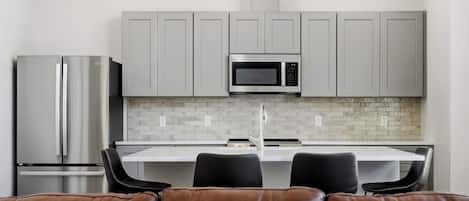Una cocina moderna con armarios grises, un refrigerador de acero inoxidable, un microondas y un lavamanos. Hay dos sillas negras en una mesa blanca.