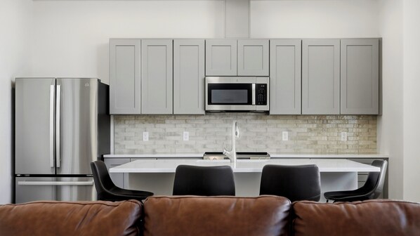 Una cocina moderna con armarios grises, un refrigerador de acero inoxidable, un microondas y un lavamanos. Hay dos sillas negras en una mesa blanca.