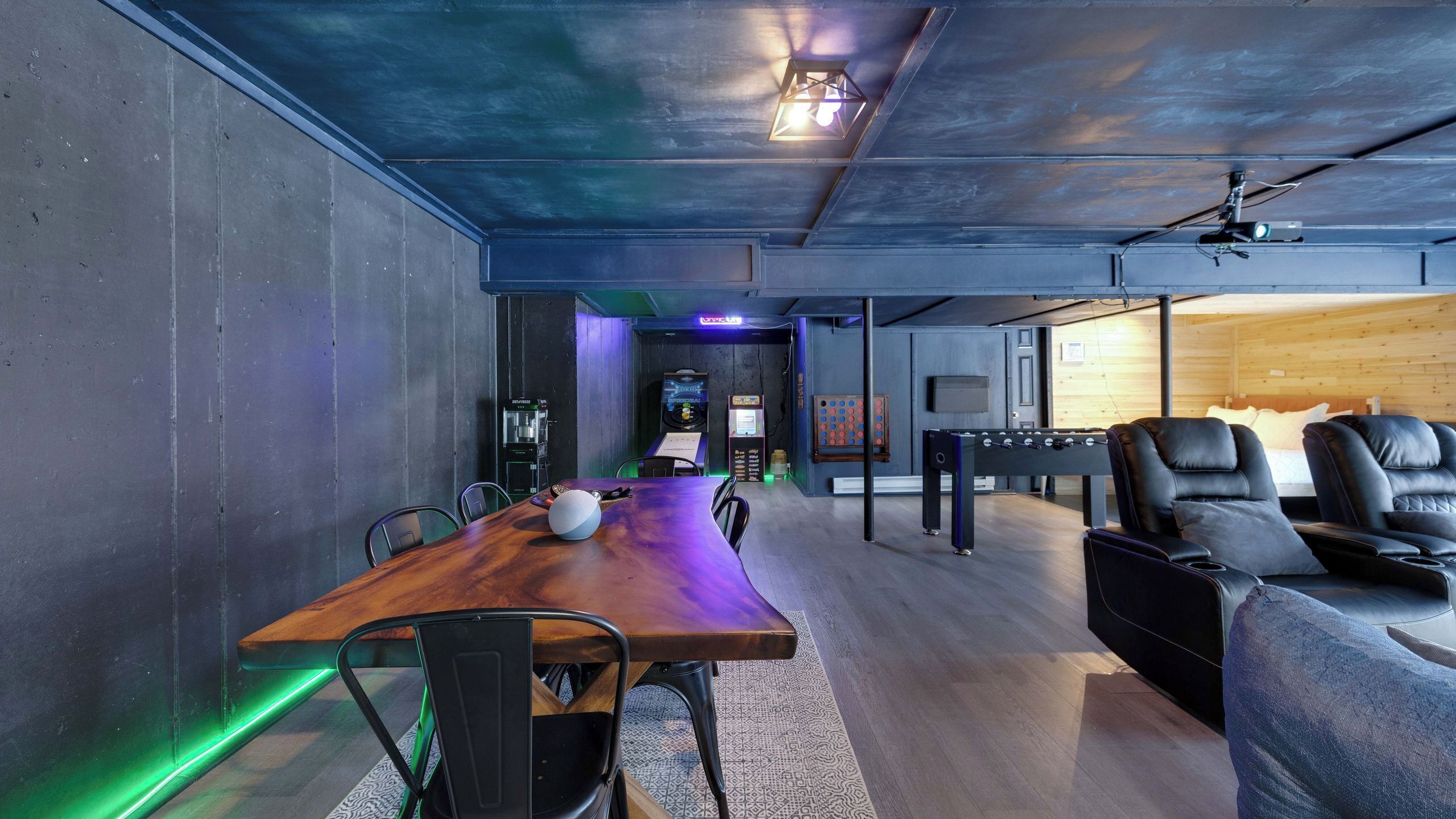 A modern room with a wooden table, chairs, a foosball table, and a gaming area with arcade machines.