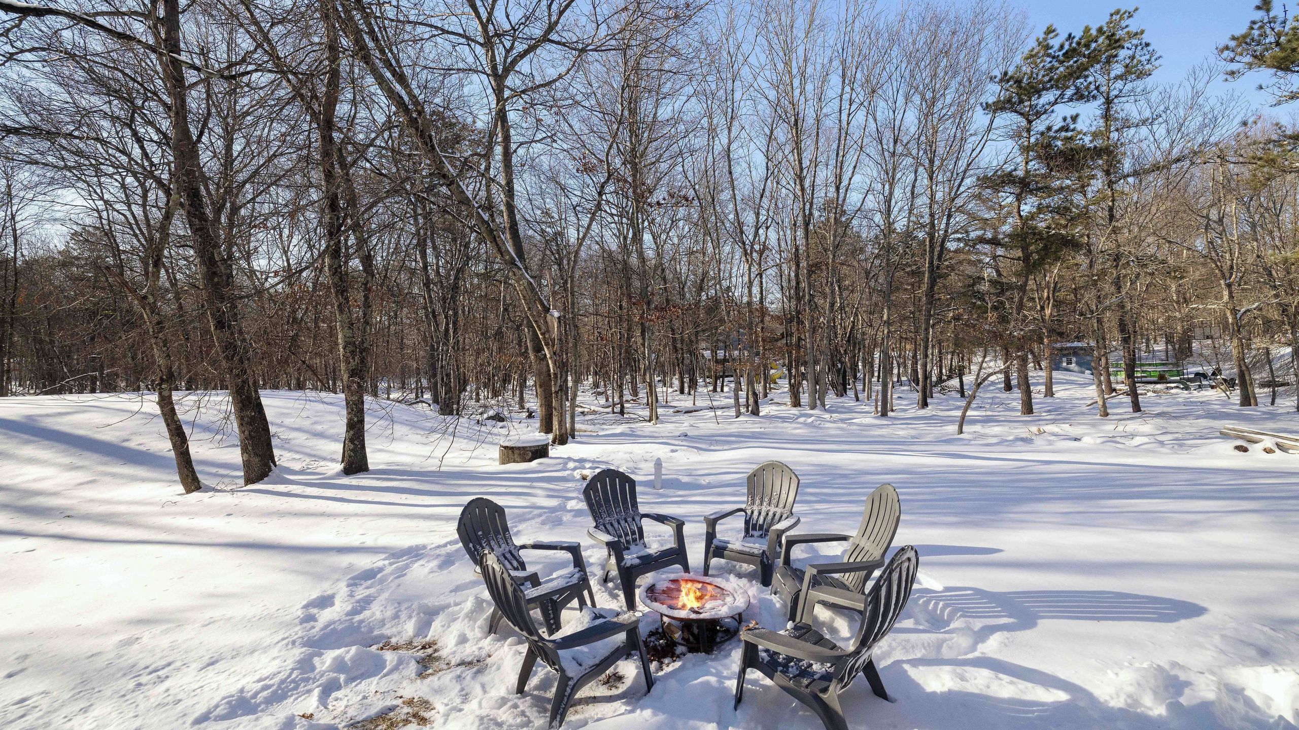 Un espacio exterior cubierto de nieve con un hogar y cuatro sillas Adirondack dispuestas a su alrededor.