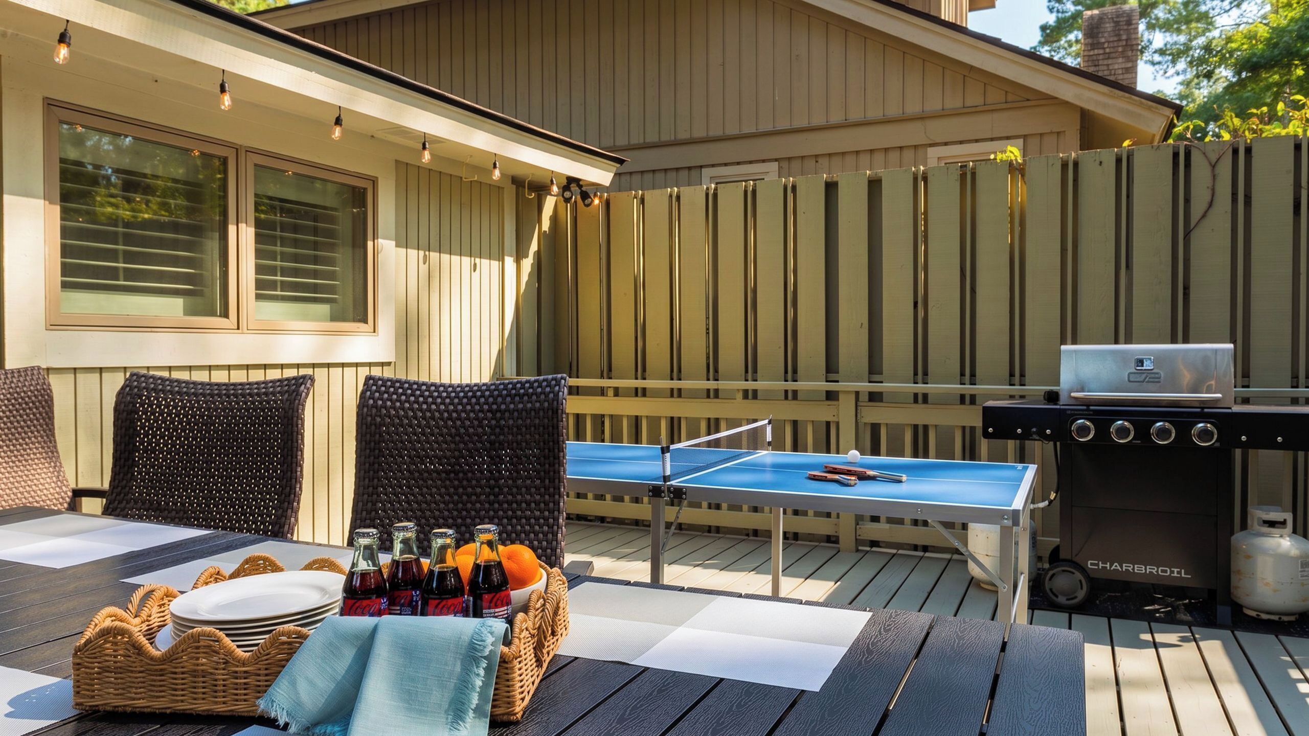 An outdoor deck with a ping pong table, wicker chairs, a grill, and a table set with plates and Coca-Cola bottles.