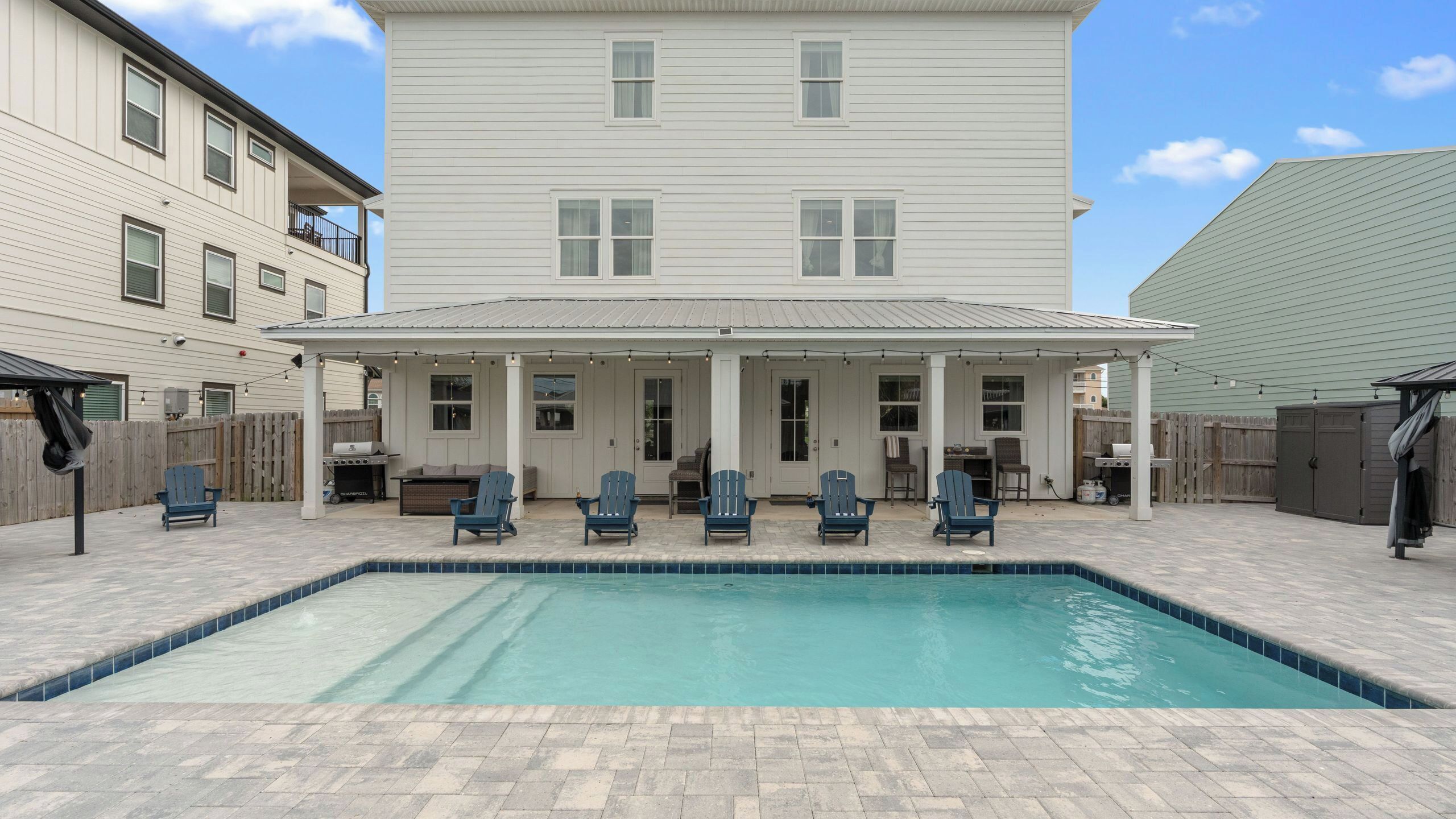 A pool area with a covered patio, outdoor seating, and a two-story building in the background.