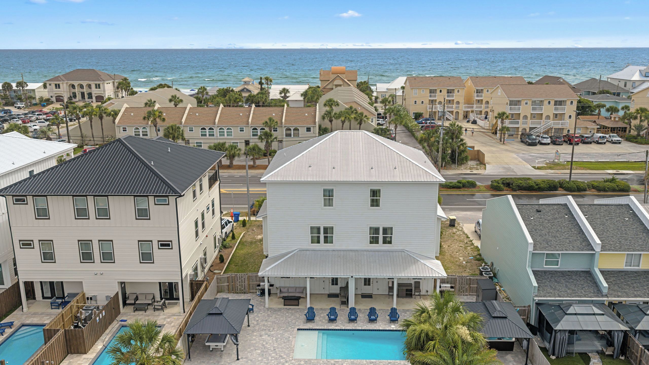 A residential area with a large white house featuring a covered patio with blue chairs, a swimming pool, and a view of the ocean.