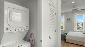 Uma lavanderia com máquina de lavar e secadora de abertura frontal, uma porta para um quarto e um ventilador de teto.