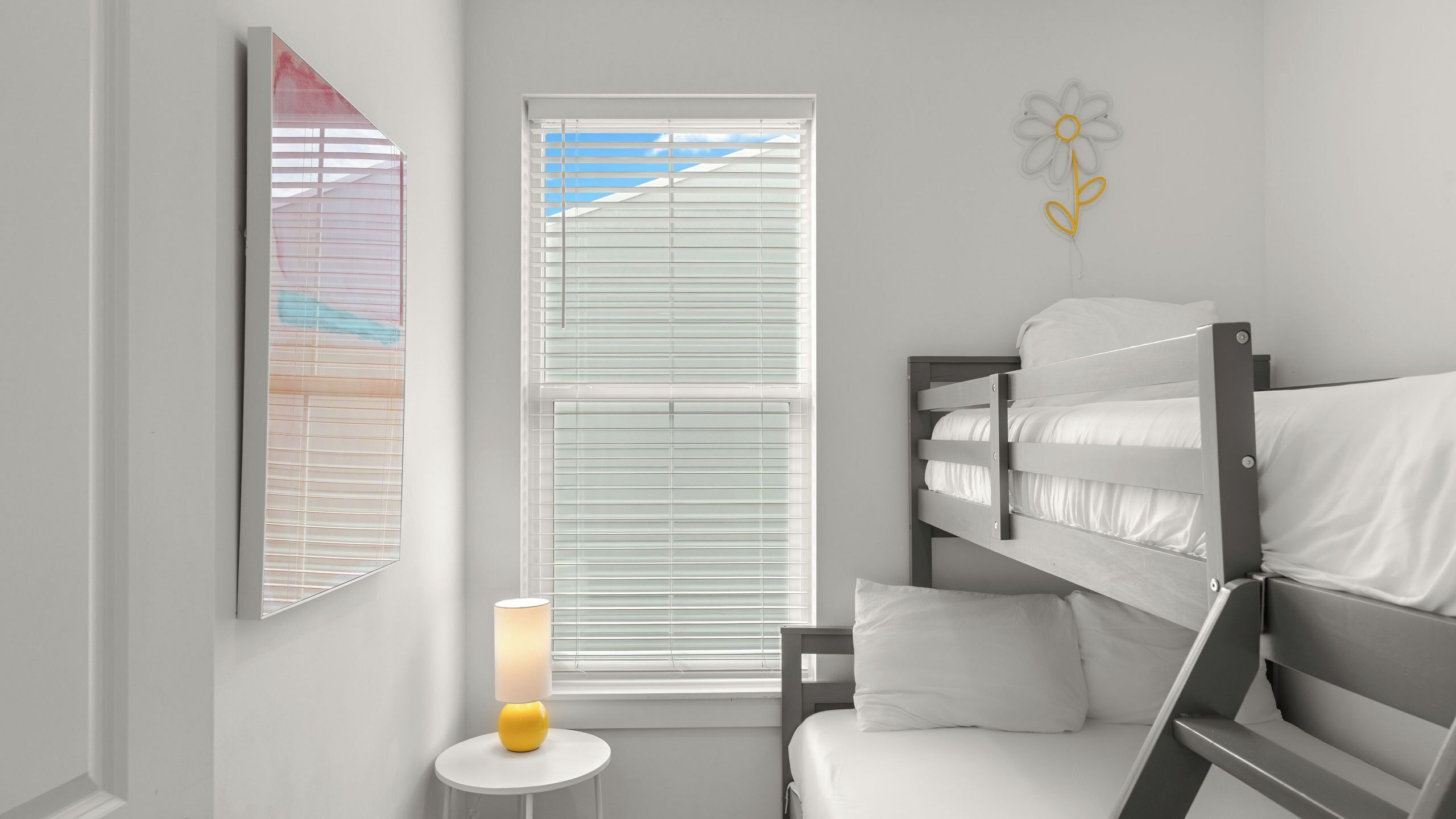 Una habitación infantil con literas, una lámpara de noche, una ventana con persianas y un adorno en la pared.