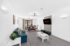 Ein Wohnzimmer mit einer Couch, einem Couchtisch, einem an der Wand befestigten Fernseher und einem Essbereich mit Tisch und Stühlen.