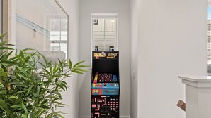 A narrow hallway with a potted plant on the left, a door with a window, and a retro arcade machine in the center. - Low Tide by Avantstay Walk to Beach Shared Pool + Hot Tub Near St. Andrews (Panama City Beach)